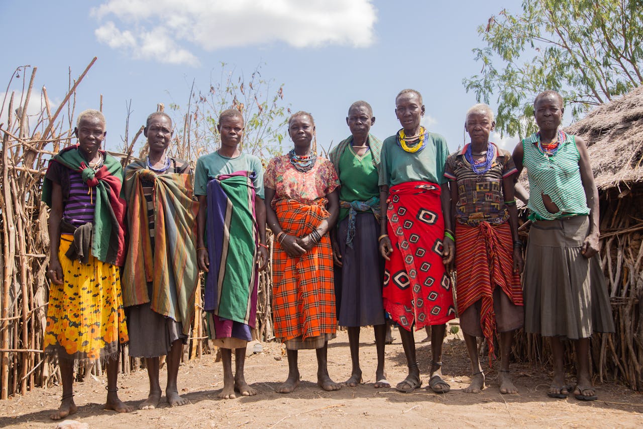 who-we-are A group of women from an African tribe in traditional attire posing outdoors in their village.